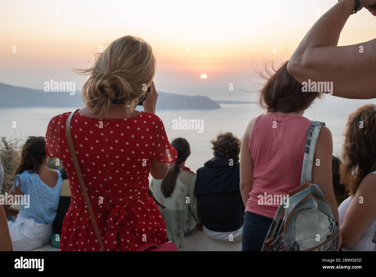 Blonde Frau in einem roten Kleid mit weißen Punkten zwischen Touristen fotografiert den atemberaubenden Sonnenuntergang von Oia in Santorini Stockfoto