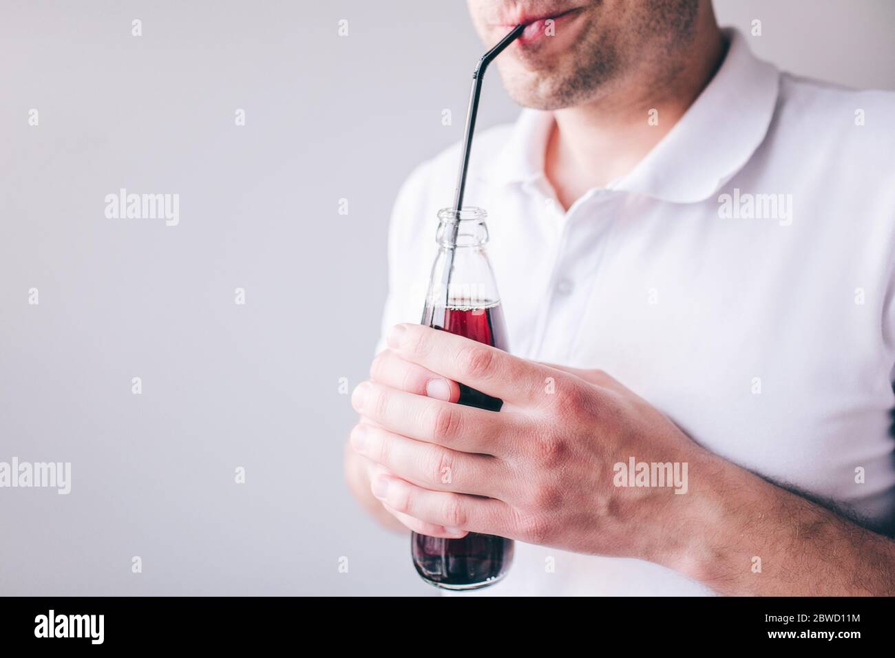 Junger Mann in weißem Hemd isoliert über Hintergrund. Schnitt Ansicht von Kerl trinken Cola durch Kunststoff Stroh. Flasche in Händen halten. Stockfoto