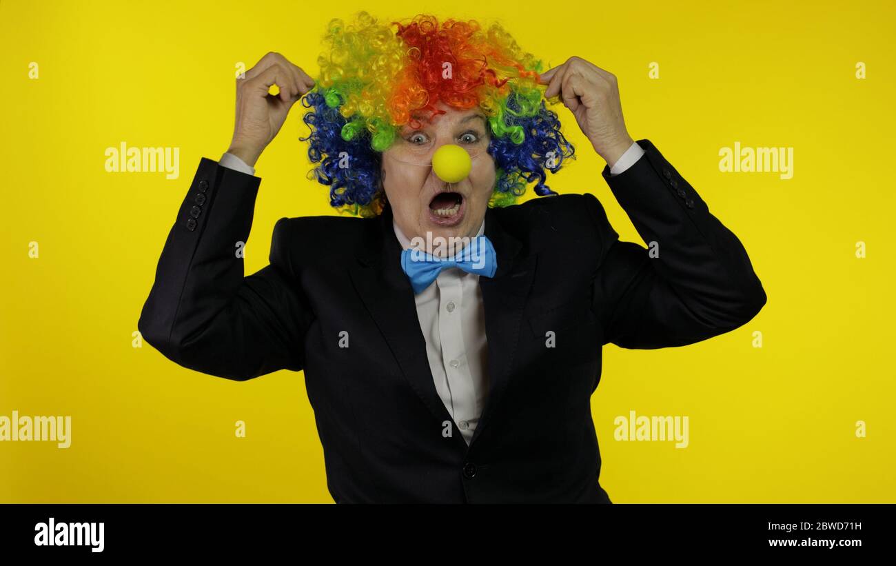 Senior kaukasischen weiblichen Clown in bunten Perücke und blaue Fliege machen dumme Gesichter, Narr um. Ältere Frau, die Spaß hat, lächelt, schaut Kamera an. Kopierbereich. Halloween. Gelber Hintergrund Stockfoto