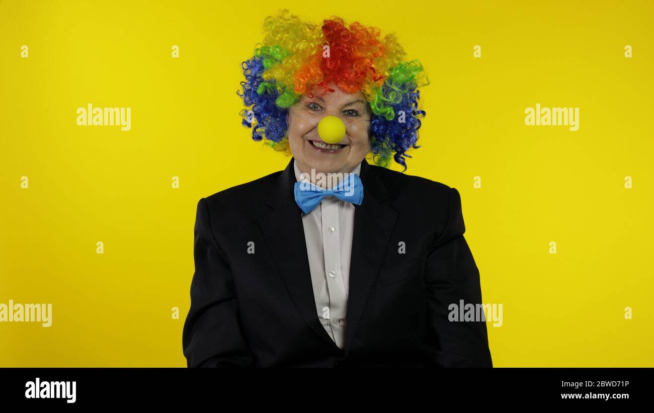 Senior kaukasischen weiblichen Clown in bunten Perücke und blaue Fliege machen dumme Gesichter, Narr um. Ältere Frau, die Spaß hat, lächelt, schaut Kamera an. Kopierbereich. Halloween. Gelber Hintergrund Stockfoto