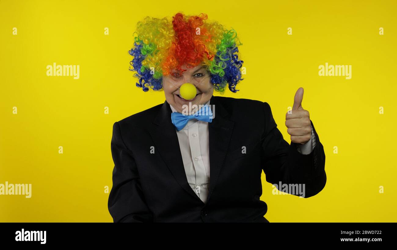 Ältere alte kaukasische Clownin in bunten Perücke und blaue Fliege zeigen Daumen nach oben. Ältere Frau, die Spaß hat, lächelnd. Kopierbereich. Halloween. Gelber Hintergrund Stockfoto
