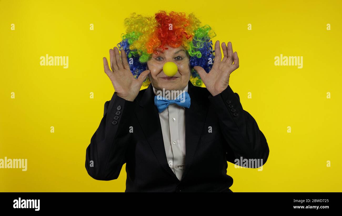 Senior kaukasischen weiblichen Clown in bunten Perücke und blaue Fliege machen dumme Gesichter, Narr um. Ältere Frau, die Spaß hat, lächelt, schaut Kamera an. Kopierbereich. Halloween. Gelber Hintergrund Stockfoto