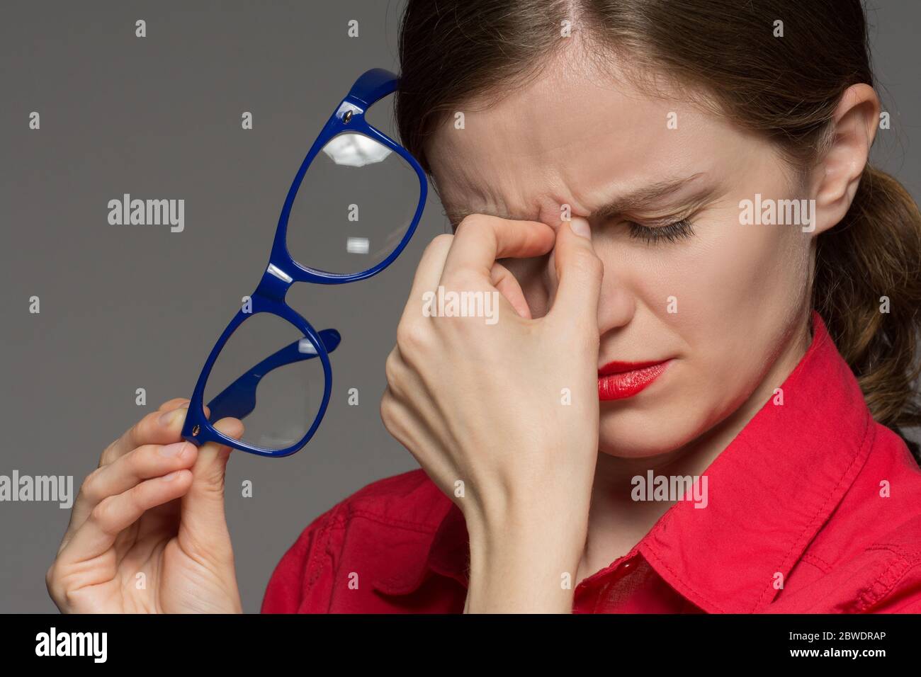 Schönes junges Mädchen in einem roten Hemd, das Wunden Augen und einen Kopf aus einer Brille auf einem grauen Hintergrund hat Stockfoto