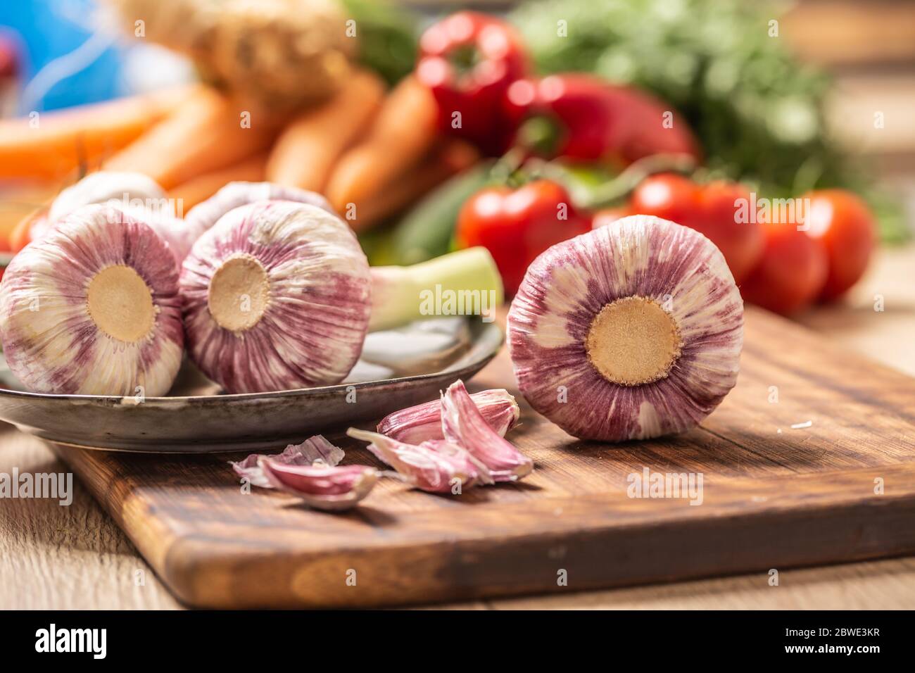 Fünf Köpfe Knoblauch auf einem Schneidebrett mit verschiedenen Gemüse im Hintergrund Stockfoto