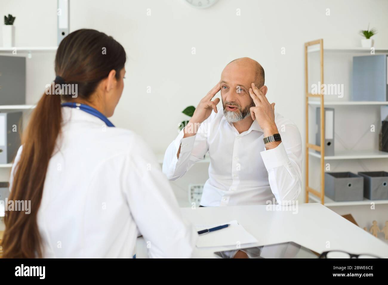 Älterer männlicher Patient mit Kopfschmerzen bei einem Arzttermin in einem weißen Büro einer medizinischen Klinik. Stockfoto
