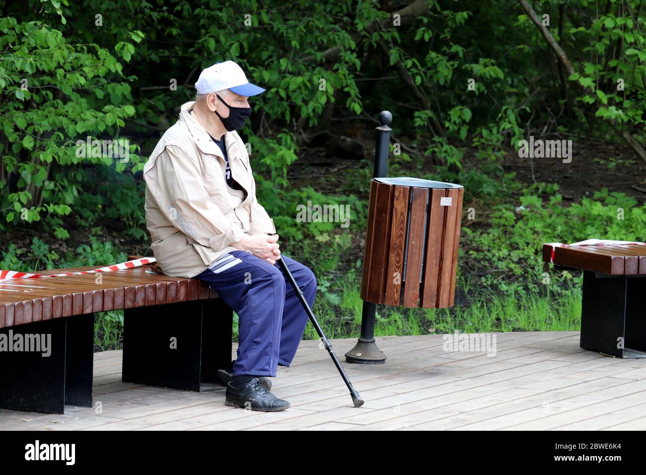 Älterer Mann in einer medizinischen Maske sitzt mit einem Stock auf einer Bank in einem Stadtpark. Alte Menschen während der Quarantäne der Covid-19-Coronavirus-Pandemie Stockfoto