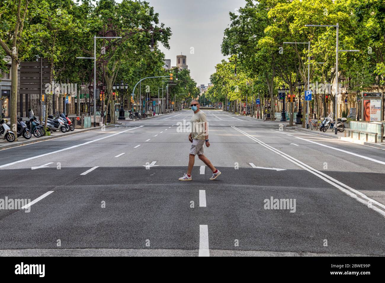 Mann, der während der Pandemie von 19 in Barcelona, Katalonien, Spanien eine Operationsmaske trägt, während er eine leere Passeig de Gracia-Allee überquert Stockfoto