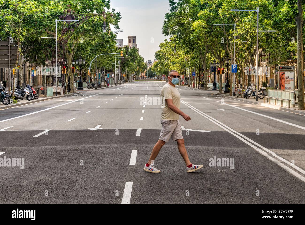 Mann, der während der Pandemie von 19 in Barcelona, Katalonien, Spanien eine Operationsmaske trägt, während er eine leere Passeig de Gracia-Allee überquert Stockfoto