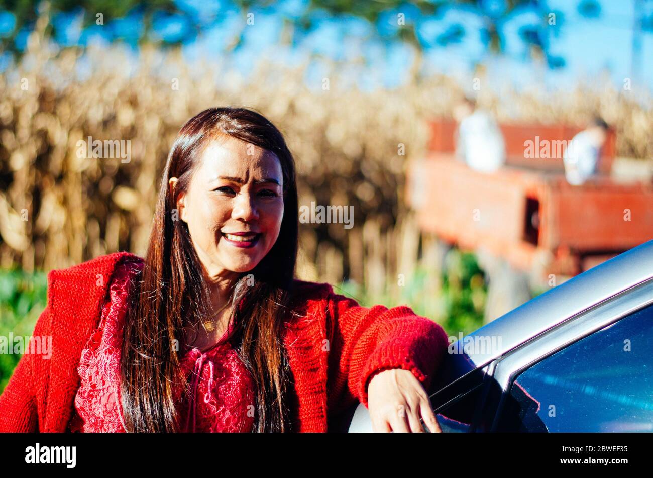 Indonesische Frau mittleren Alters in roten Kleidern posiert und lächelt vor einem blauen alten LKW von einem Bauernhof. Stockfoto
