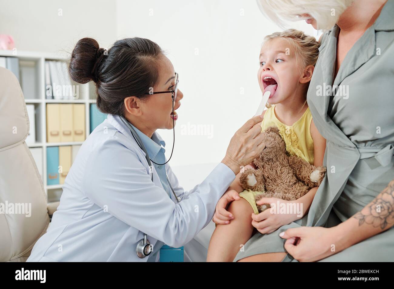 Doktor bittet kleines Mädchen, den Mund weit zu öffnen und Spatel zu verwenden, wenn sie ihren Hals überprüft Stockfoto