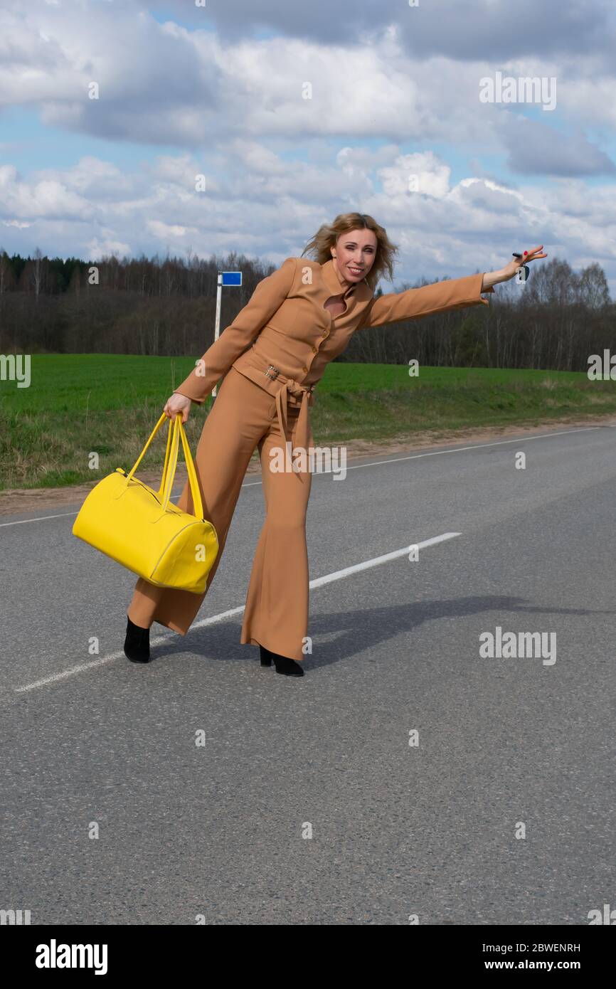 Elegante Frau mittleren Alters in einem modischen Hosenanzug und mit einer gelben Reisetasche in der Hand überquert die Straße. Das Konzept der Erholung von der Stadt und Stockfoto