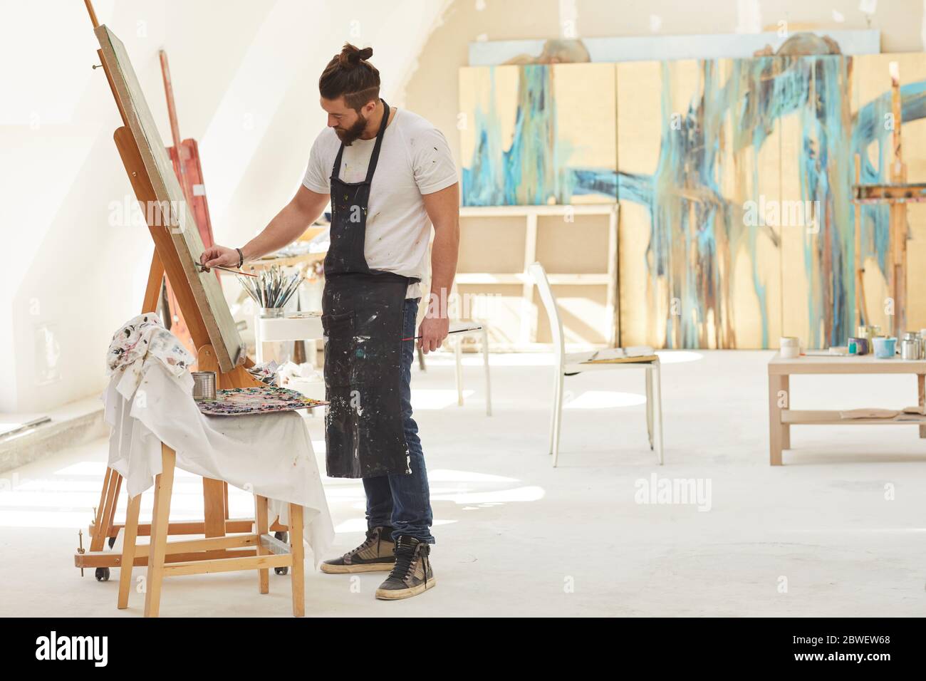 Weitwinkel-Seitenansicht auf zeitgenössische bärtige Künstler Gemälde Bild auf Staffelei während der Arbeit in geräumigen Kunststudio durch Sonnenlicht beleuchtet, Kopierraum Stockfoto