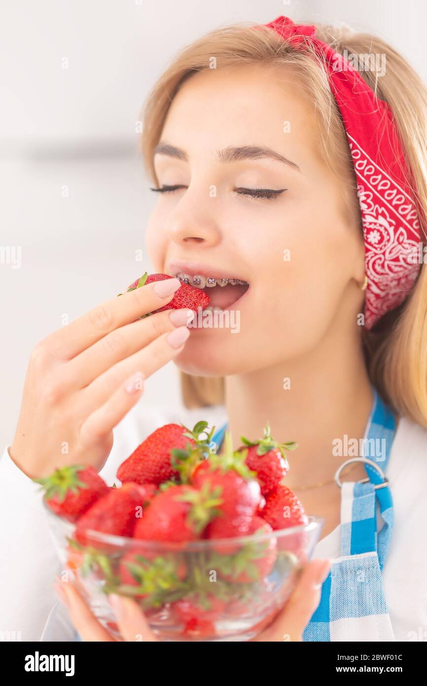 Schöne junge Frau, die den Geschmack eines Bisses von einer frischen Erdbeere mit einer Schüssel mit mehr Früchten vor sich genießt. Stockfoto