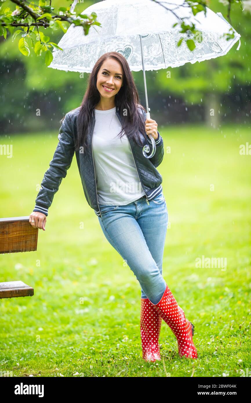 Lächelnd gut aussehende Frau lehnt sich gegen eine Parkbank unter dem Regenschirm geschützt. Stockfoto