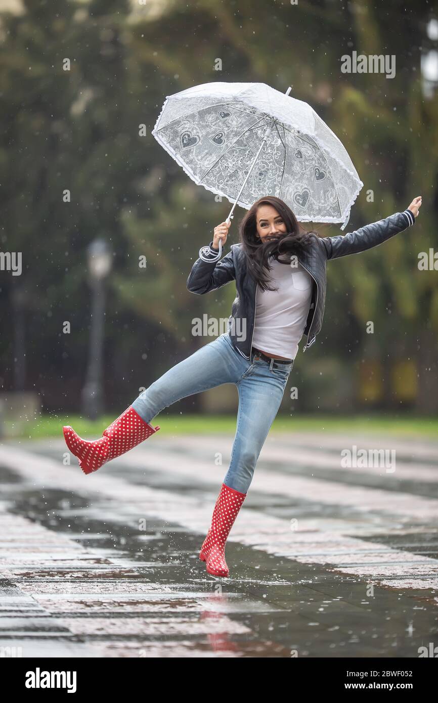 Die Frau springt an einem regnerischen Tag mit Regenschirm und roten Polka Dot Regenstiefeln vor Freude. Stockfoto