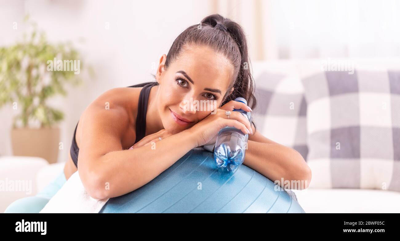 Porträt einer schönen Frau, die sich an beiden Ellbogen auf einen fittierten Ball stützt und Wasser in Flaschen trinkt. Stockfoto