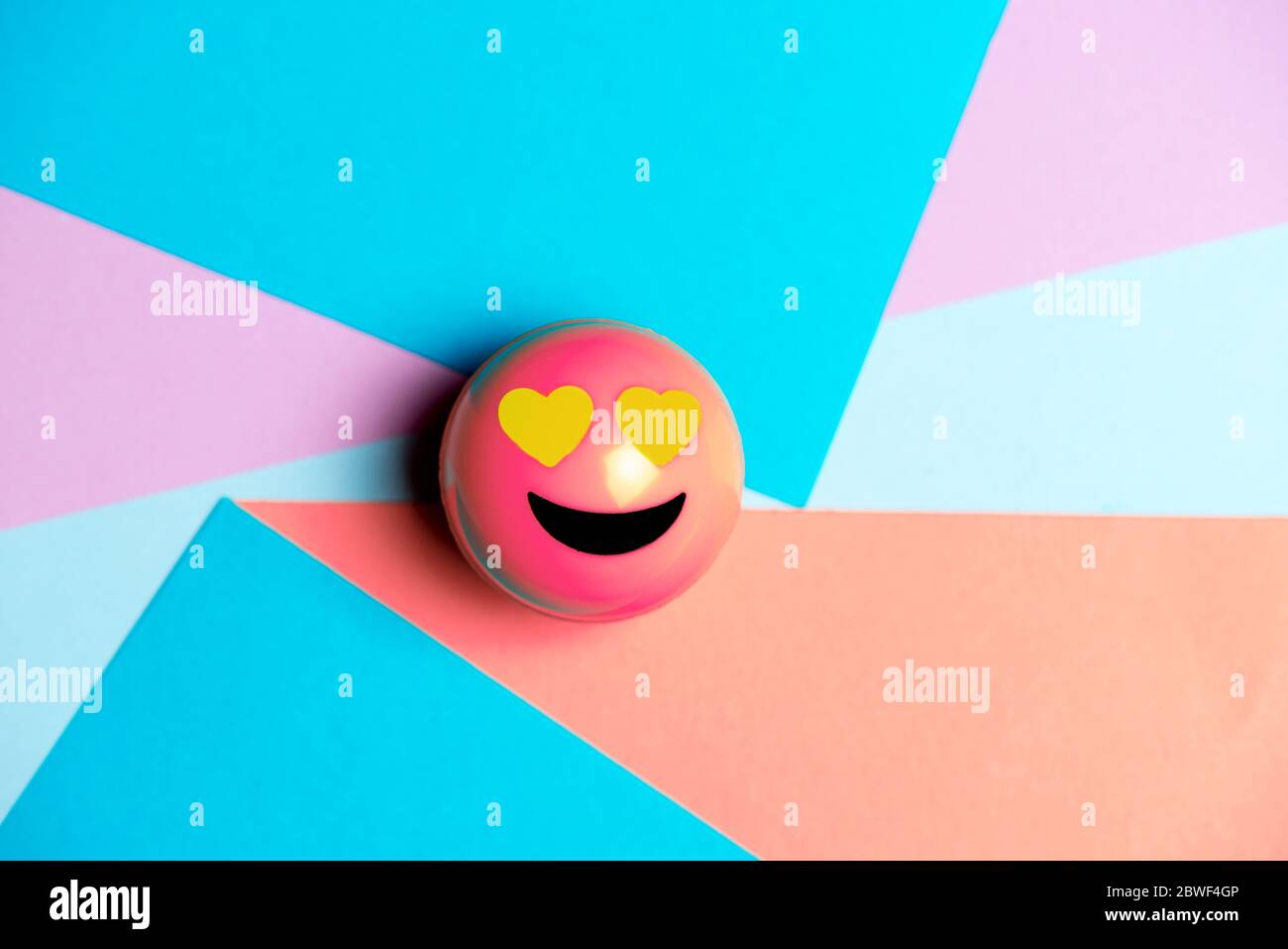 Lächelndes rosa Emoticon mit Herzaugen, auf mehrfarbige geometrische Textur, Social-Media-Konzept Stockfoto