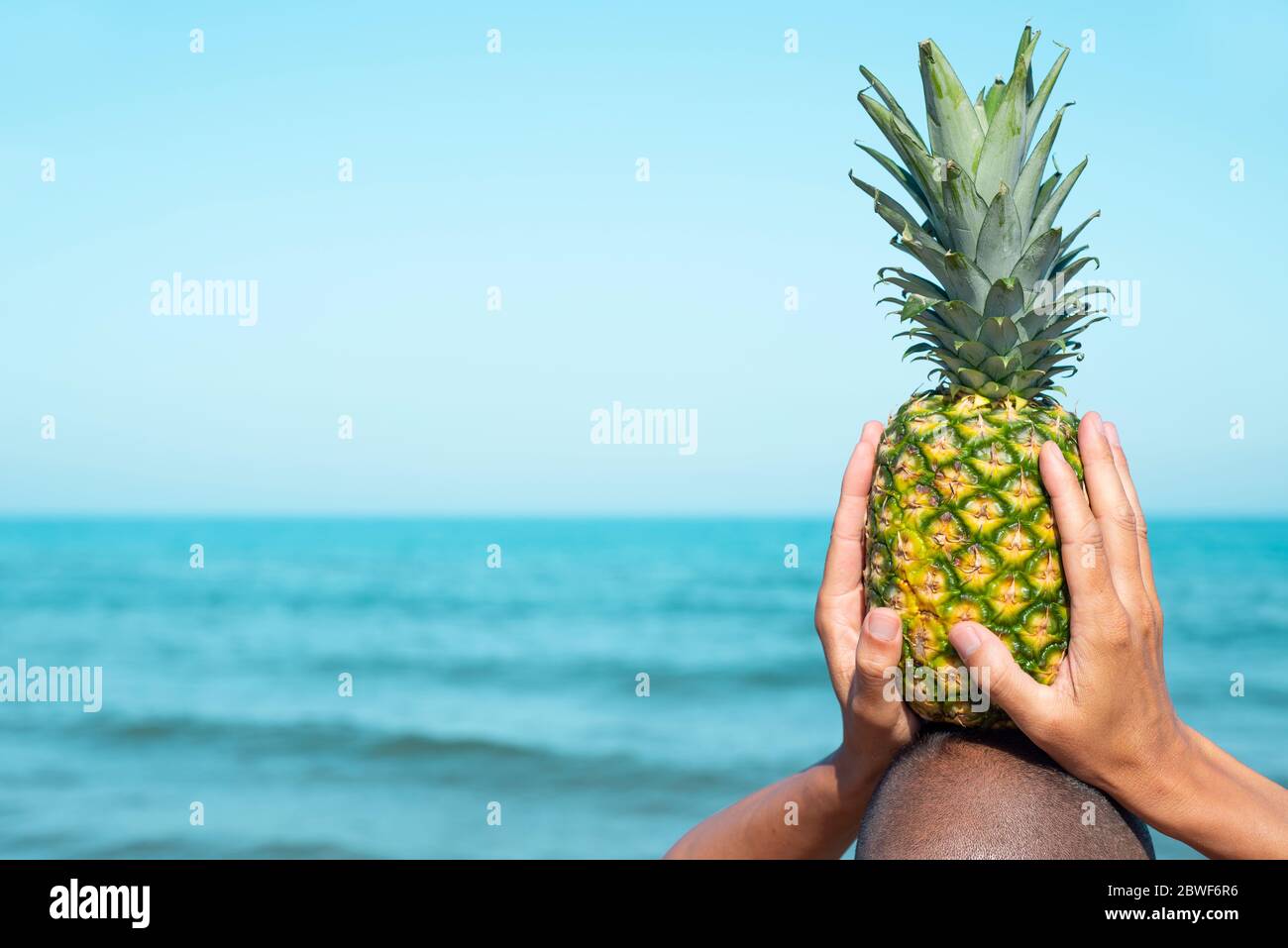 Nahaufnahme eines Mannes, der von hinten am Strand vor dem Meer gesehen wird und eine Ananas auf dem Kopf hält, mit einer leeren Fläche auf der linken Seite Stockfoto