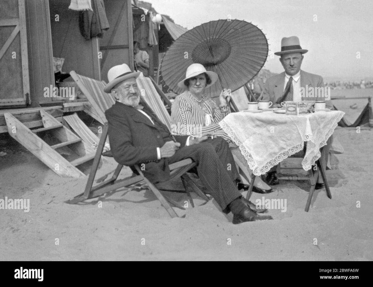 Am Meer genießen drei Menschen, formell gekleidet und mit Hüten, Umbauten im Freien, UK um 1930. Sie sitzen auf Liegestühlen vor einer Strandhütte. Auf dem Picknicktisch liegen Teebecher und eine Schachtel Kekse. Stockfoto