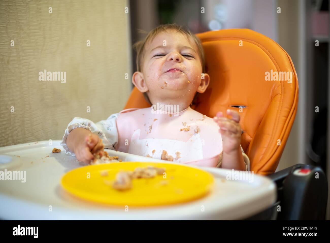 Problem beim Füttern des Babys. Niedliches Baby in einem orangefarbenen Kindersitz schaut auf die Kamera und grinst. Krümel und eine orangefarbene Platte auf dem Tisch. Nahaufnahme, Vorderansicht Stockfoto
