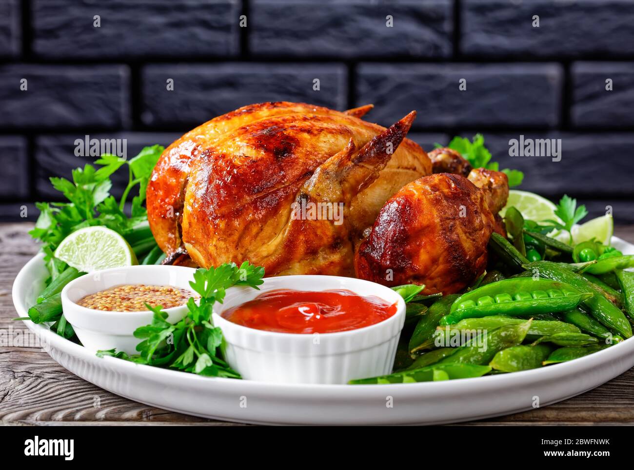 Hähnchenbraten mit gebratenen Schoten von grünen Erbsen, frischen grünen Zwiebeln, Petersilie, Limette, Tomatensauce und Senf auf einem weißen Teller auf einem dunklen w Stockfoto