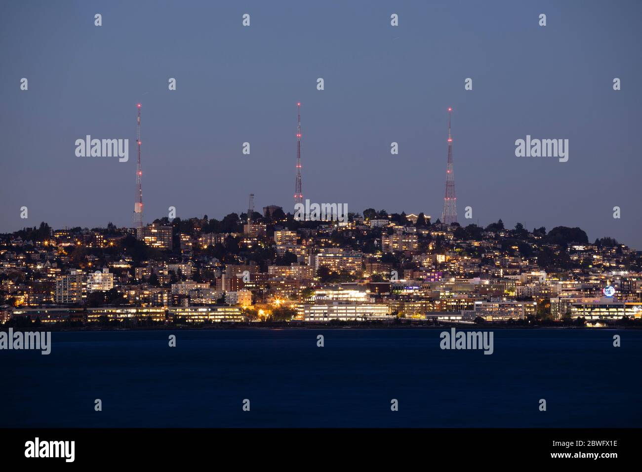 Beleuchtete Stadtgebäude am Wasser bei Nacht mit drei Kommunikationstürmen im Hintergrund, Seattle, Washington, USA Stockfoto