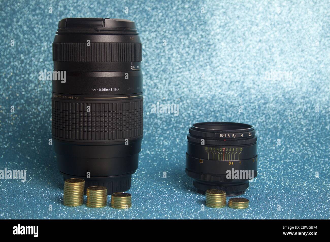 Billiges und altes Objektiv mit manueller Bedienung im Vergleich zu teurem Objektiv mit Autofokus auf blauem Hintergrund mit Münzen, die den Wert der Objektive kennzeichnen. Stockfoto