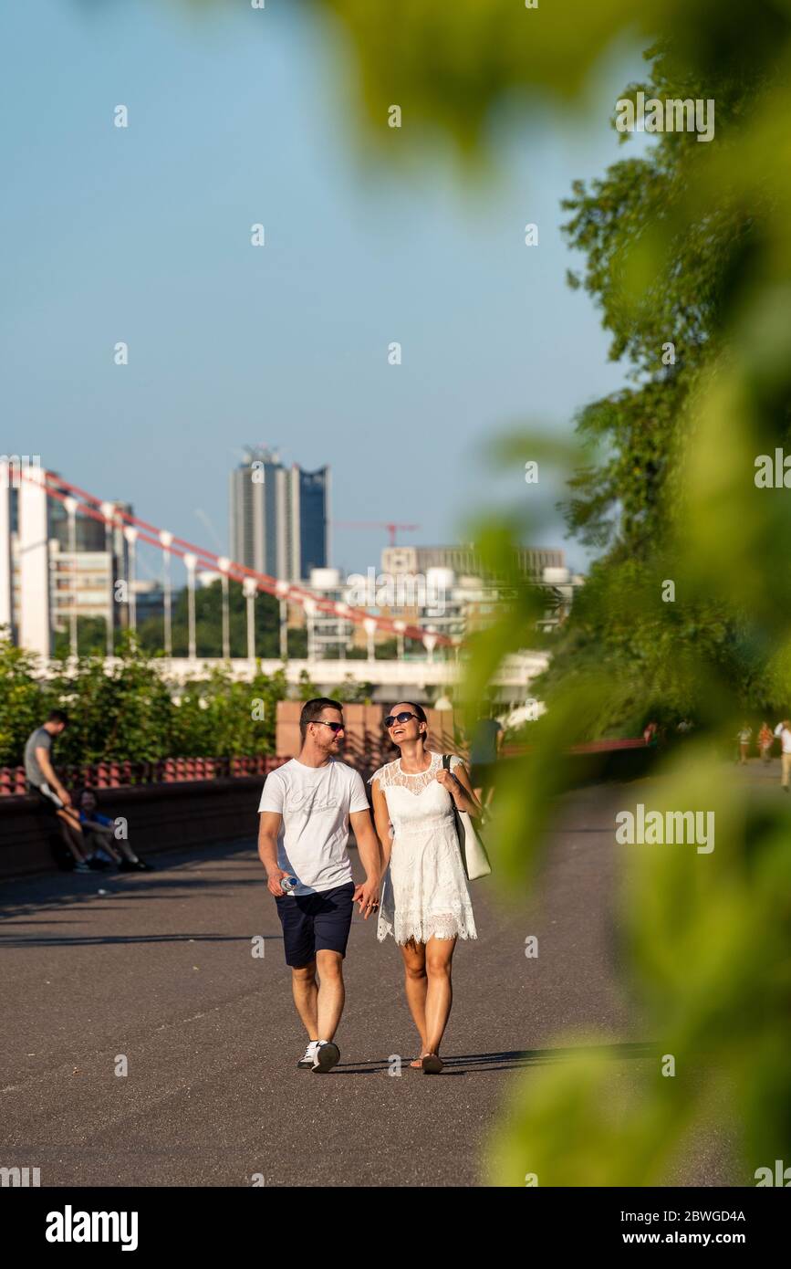 Ein trendiges junges Paar geht an einem warmen, sonnigen Sommertag auf dem themspfad im Battersea Park Chelsea London an den Händen Stockfoto
