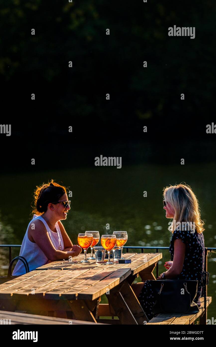 Zwei Frauen genießen während der goldenen Stunde Getränke bei Sonnenuntergang am Wasser im Battersea Park, London, mit Aperol Spritz und warmem Abendlicht. Stockfoto