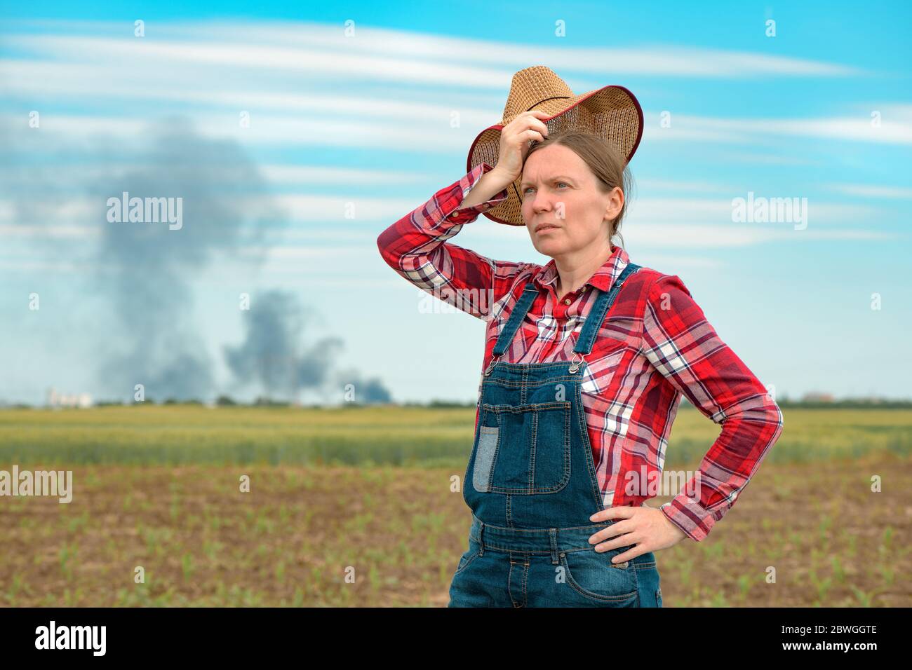 Besorgte Farmerin in Maisfeld Blick auf schwarzen Rauch am Horizont, Konzept der Versicherung in der Landwirtschaft und Landwirtschaft Stockfoto