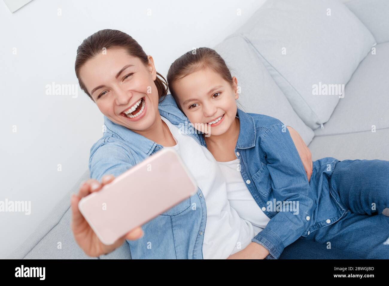 Junge Erwachsene Frau und ihre kleine Tochter sitzen zusammen auf der Couch und machen Selfie Stockfoto