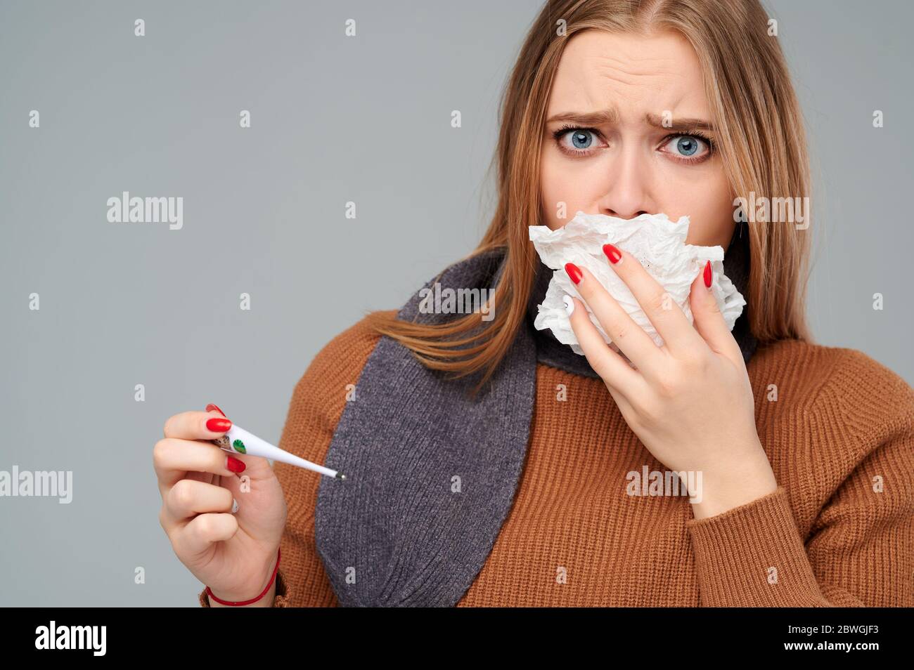 Frau im Pullover und Schal husten in eine Serviette und messen die Temperatur Stockfoto