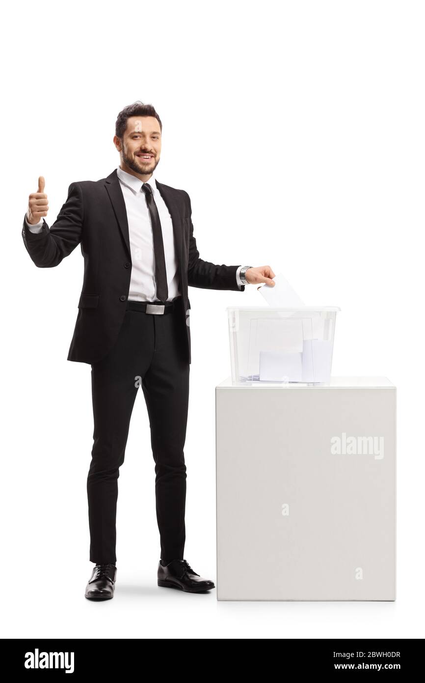 In voller Länge Porträt eines Geschäftsmannes in einem Anzug, der eine Stimme in einer Wahlurne abgibt und die Daumen nach oben isoliert auf weißem Hintergrund zeigt Stockfoto
