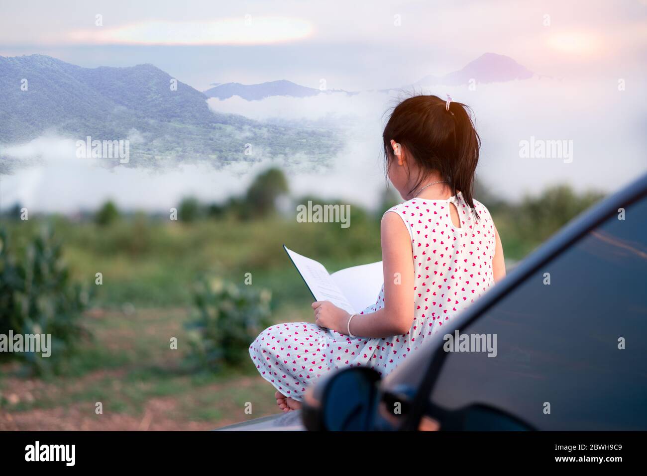 Kleines Mädchen sitzen und entspannen auf einem Auto Lesen eines Buches in einer grünen Natur Hintergrund Stockfoto