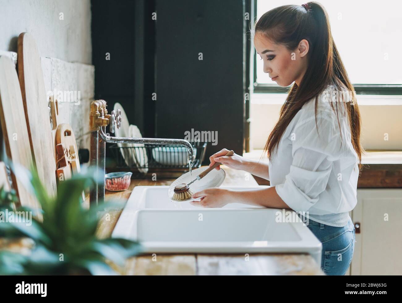 Junge Frau wäscht Geschirr mit Holzbürste mit Naturborsten am Fenster in der Küche. Konzept ohne Verschwendung Stockfoto