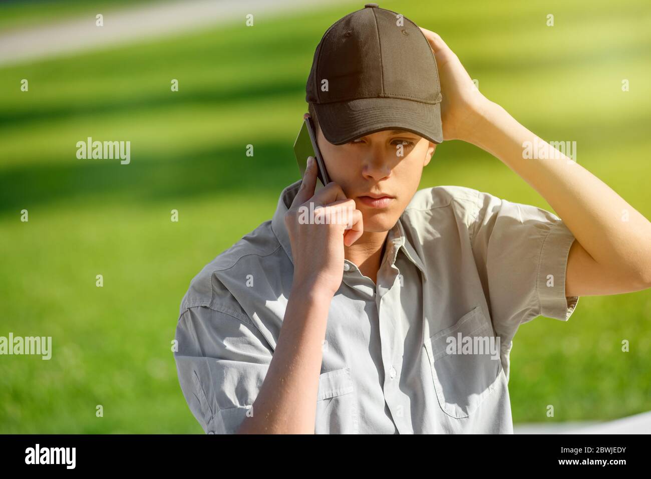 Junger Teenager, der eine Kappe trägt, die draußen vor einem grünen Gras steht und auf seinem Handy plaudert Stockfoto