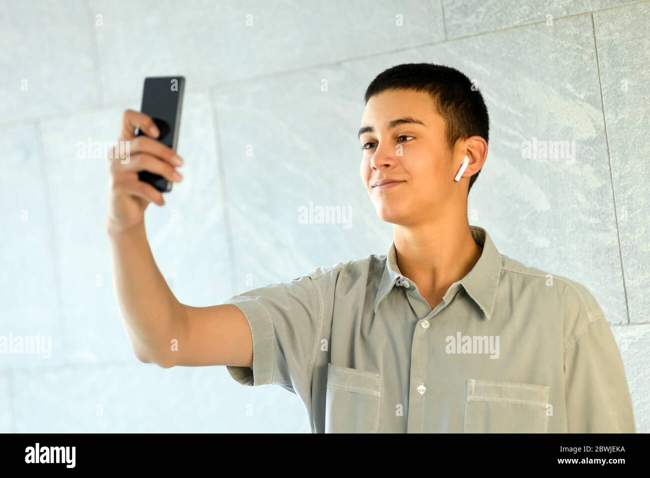 Junger Mann mit Ohrstöpsel, der ein Selfie auf seinem Handy macht, während er lächelnd für die Kamera gegen eine weiße kommerzielle Wand posiert Stockfoto