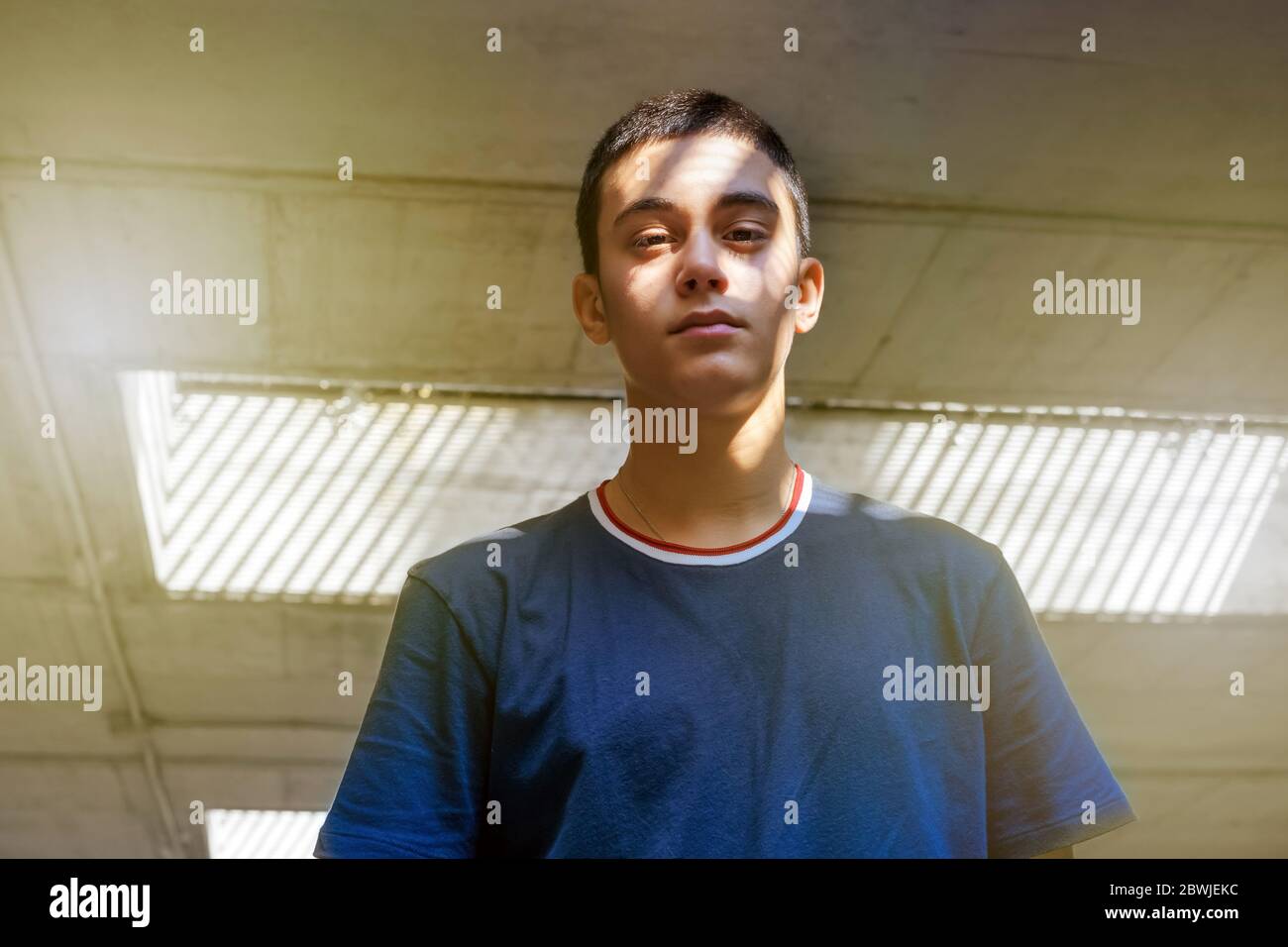 Teenager posiert mit Licht und Schatten auf seinem Gesicht in einem niedrigen Winkel, während er mit nachdenklichem Ausdruck auf die Kamera herabblickt Stockfoto