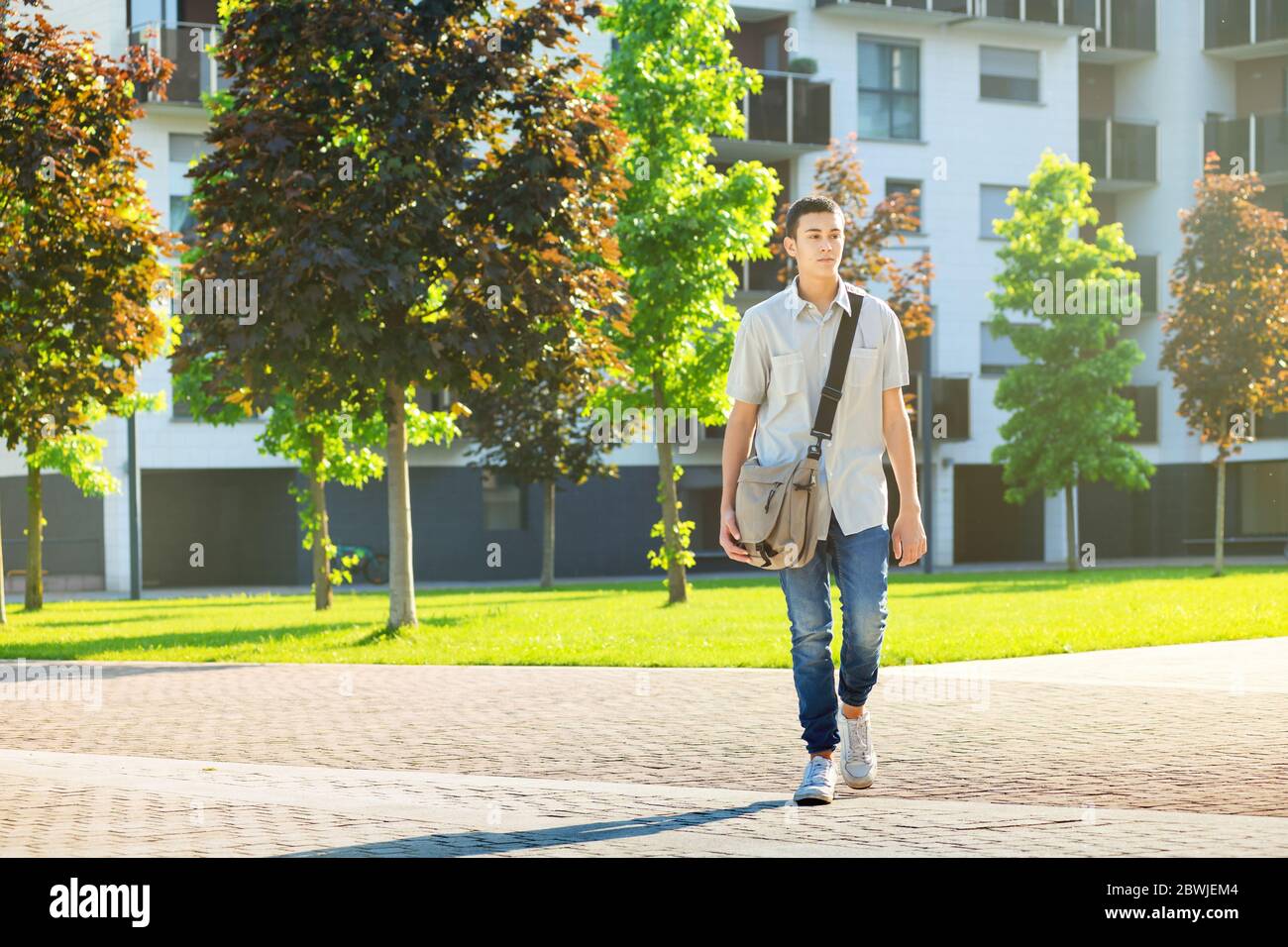 Junger Mann, der mit einer Tasche Bücher über die Schulter auf die Uni oder Universität aufbricht, während er in einem städtischen Komplex über eine gepflasterte Straße geht Stockfoto