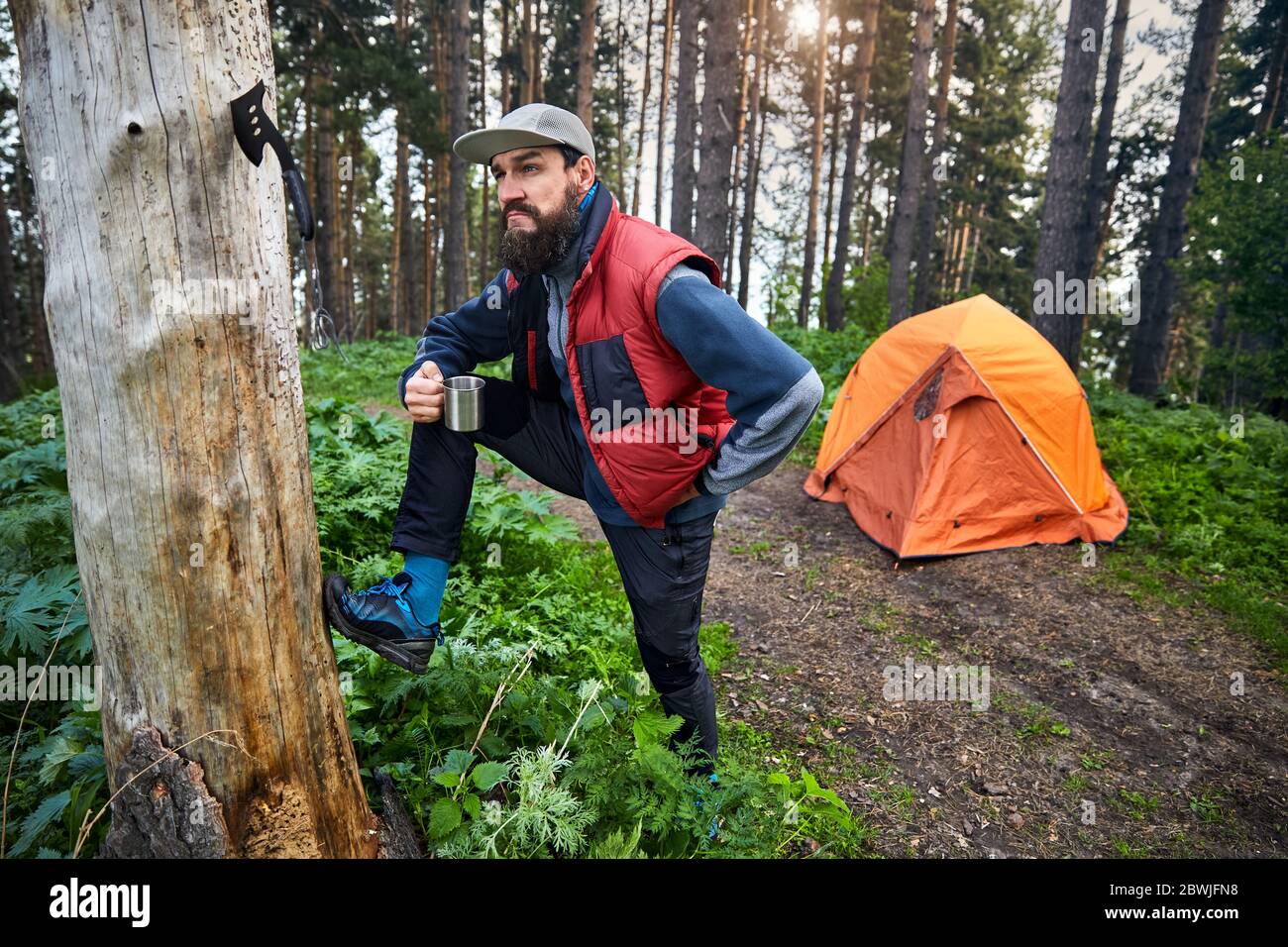 Bärtiger lustiger Mann mit Tasse Kaffee in der Nähe von orangen Zelt im Bergwald draußen Stockfoto