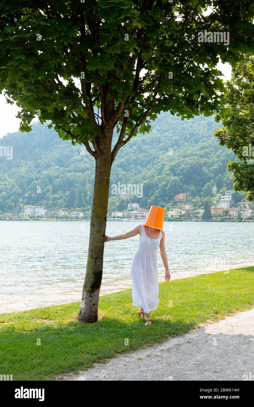Junge Frau am See mit einem orangefarbenen Eimer auf dem Kopf und einem langen Kleid und weißem Rock Stockfoto