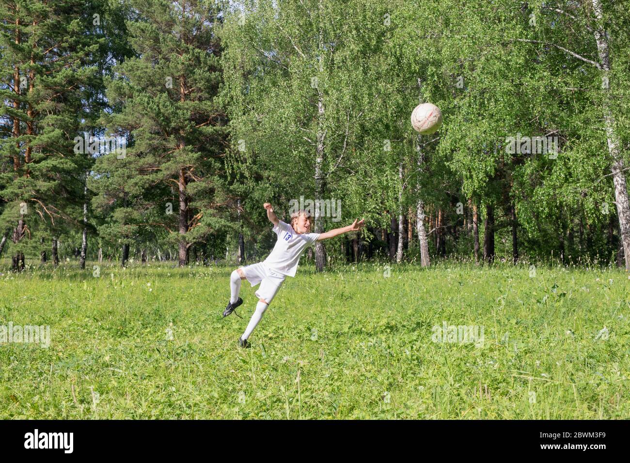 Junge in weißer Fußballuniform Fußball spielen im grünen Sommerpark auf Waldhintergrund Stockfoto