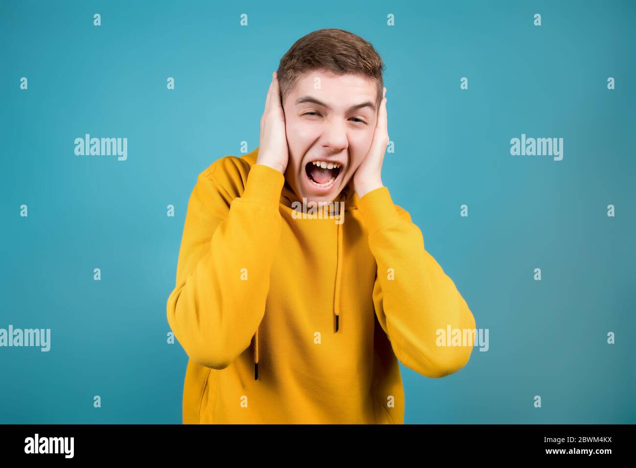 Teenager in einem gelben Sweatshirt auf blauem Hintergrund schreit und hält seinen Kopf in den Händen Stockfoto