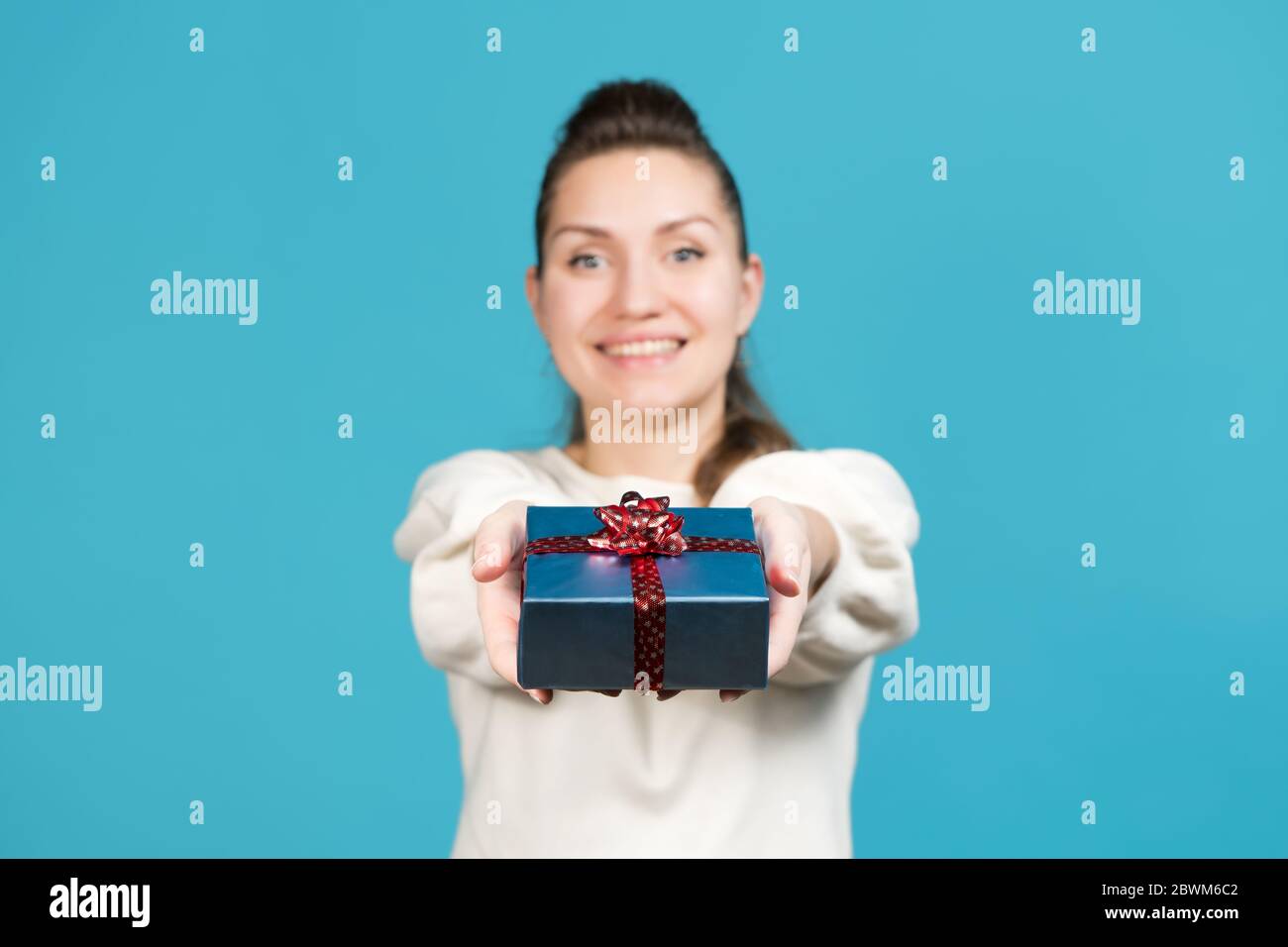 Mädchen hält ein kleines Geschenk und schaut direkt auf die Kamera. Mädchen in Unschärfe, mit Schwerpunkt auf das Geschenk. Blauer Hintergrund, Studio Stockfoto