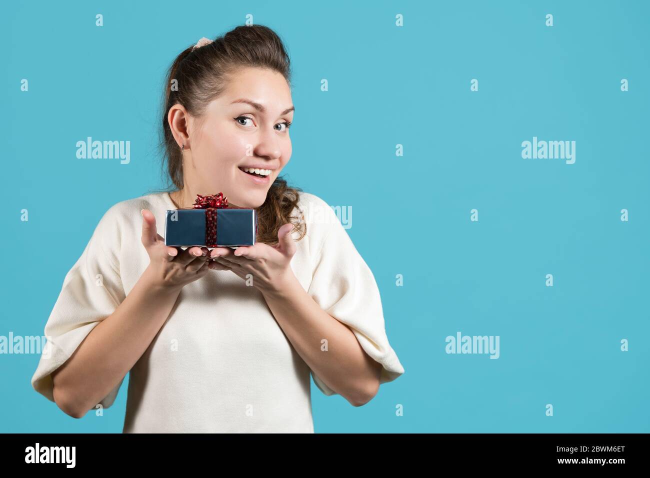 Junge Frau hält ein kleines Geschenk auf ihren Handflächen nahe bei sich und lächelt hell. Blauer Hintergrund, Kopierbereich Stockfoto