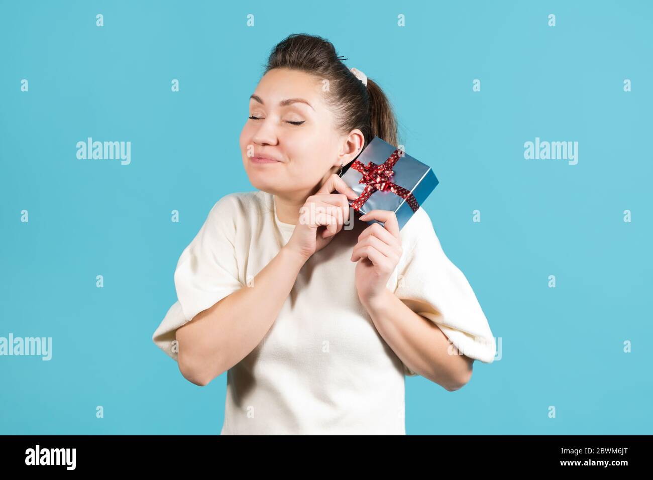 Junge Frau freut sich über ein Geschenk, bringt es ihr ins Gesicht und lächelt. Blauer Hintergrund Stockfoto