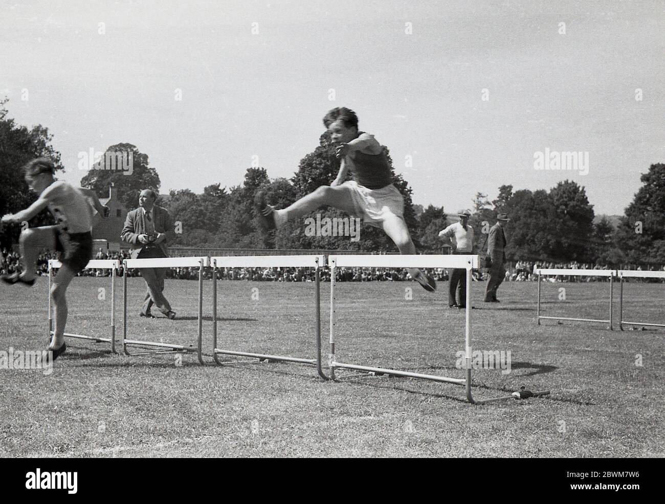 1950er Jahre, historisch, Sommerzeit und ein Schulsporttag, Schüler in einem Hürdenlauf auf einer Grasbahn in einem Sportplatz konkurrieren, Devon, England, Großbritannien, mit einem Jungen zeigt gute Technik über die Barrieren. Hürdenlauf ist eine Sportart, die sowohl Laufen und Springen über Hindernisse mit Geschwindigkeit und so ist eine technisch schwieriger athletischen Veranstaltung, die einfach schnell in einer geraden Linie laufen. Stockfoto