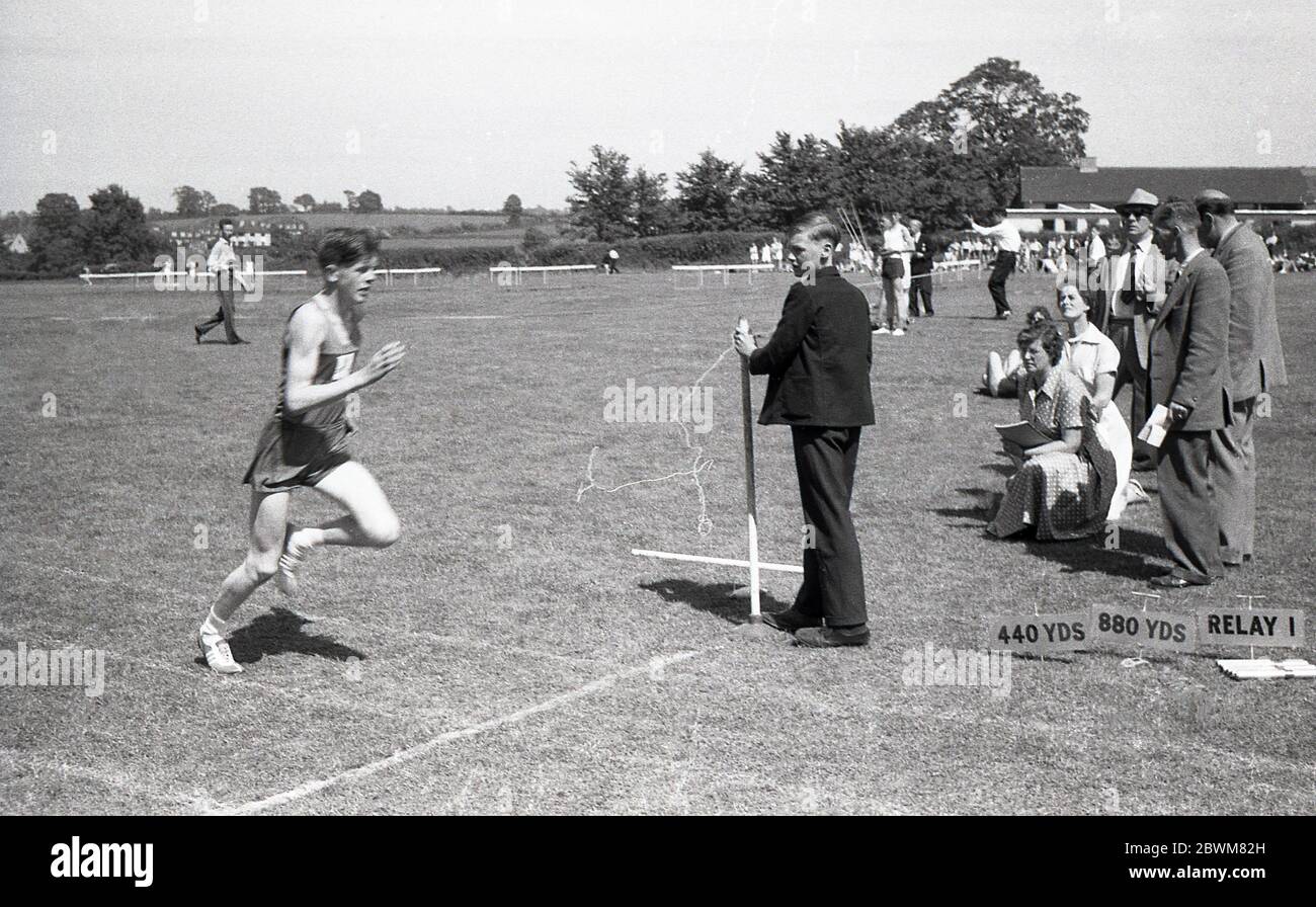 1950er Jahre, historisch, Sommerzeit und ein Schulsportwettbewerb, draußen auf einem großen Sportplatz, ein älterer Junge in einem Rennen auf einer Grasbahn, beobachtet von Lehrern und Beamten, Devon, England, Großbritannien. Stockfoto
