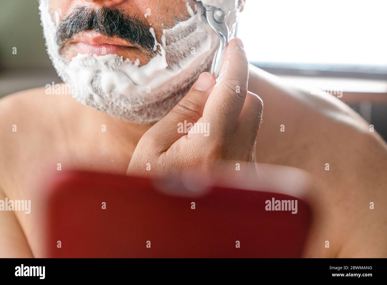 Porträt eines beschäftigten und abgelenkten jungen Mannes, der sich während der SMS auf seinem Smartphone den Bart rasieren lässt. Der Mensch nutzt das Smartphone als Spiegel, wenn er rasiert. Stockfoto