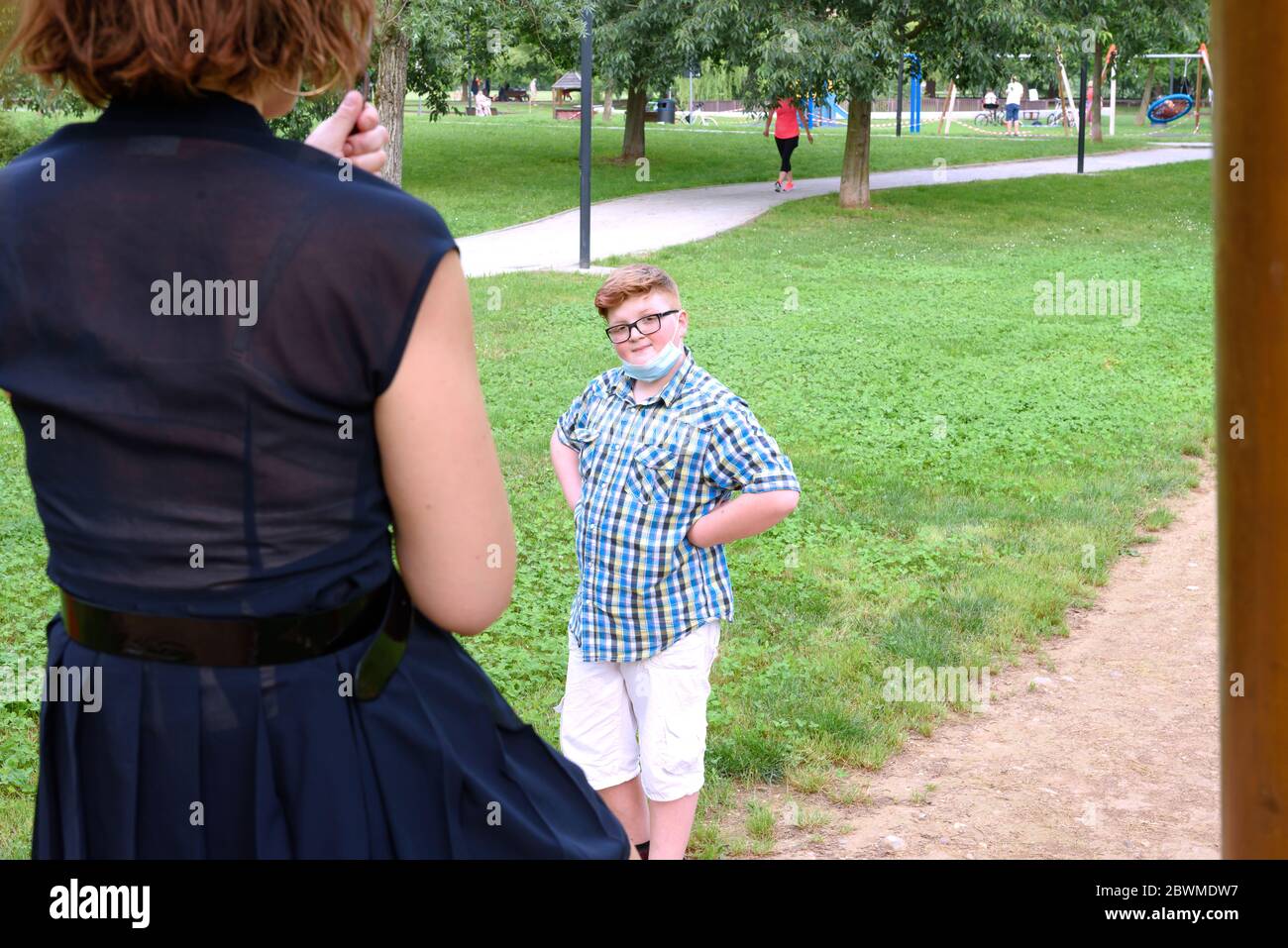 Junge, der Brille und Maske trägt, in einem karierten Hemd gekleidet, schaut auf ein Mädchen auf dem Spielplatz. Kind vor Mädchen in einem öffentlichen Park. Stockfoto